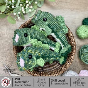 May include: Three handmade crochet fish in a woven basket. The fish are primarily green with black eyes and white and pink accents. The basket is brown, and the background includes greenery and yarn. The fish are a unique handmade item.