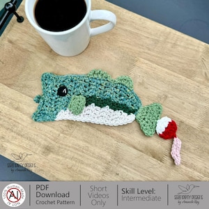 May include: A handmade crochet alligator drinks coaster in shades of blue, green, and white, with a black button eye and a fishing bobber attached. The coaster is placed on a wooden surface next to a white mug of coffee. The text "SERET DIPITY DESIGNS by Amanda Ray" is visible.