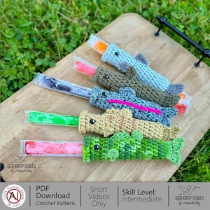 May include: Crocheted popsicle holders in the shape of various sea creatures, including a shark and a crocodile. The holders are in shades of blue, green, and tan, with popsicles inside. The image also includes text that says "SERENDIPITY DESIGNS by Amanda Ray".