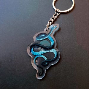 Peut inclure: Un porte-clés en acrylique transparent avec un serpent bleu et noir enroulé sur lui-même. Le porte-clés est doté d'un anneau et d'une chaîne en métal argenté.