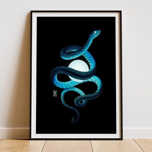 Art print of a coiled blue snake wrapped around a glowing orb of light. The background is solid black, creating a dramatic contrast with the snake and the soft glow from the orb. The print is framed in a thin black frame. The frame is placed on a light wooden floor rested against a white wall.