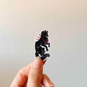 Puede incluir: Pegatina de lobo negro con detalles en rojo. El lobo se representa en una pose din&aacute;mica, con l&iacute;neas rojas que irradian de su cuerpo. La pegatina est&aacute; sujeta por una mano sobre un fondo neutro. Ilustraci&oacute;n gr&aacute;fica.