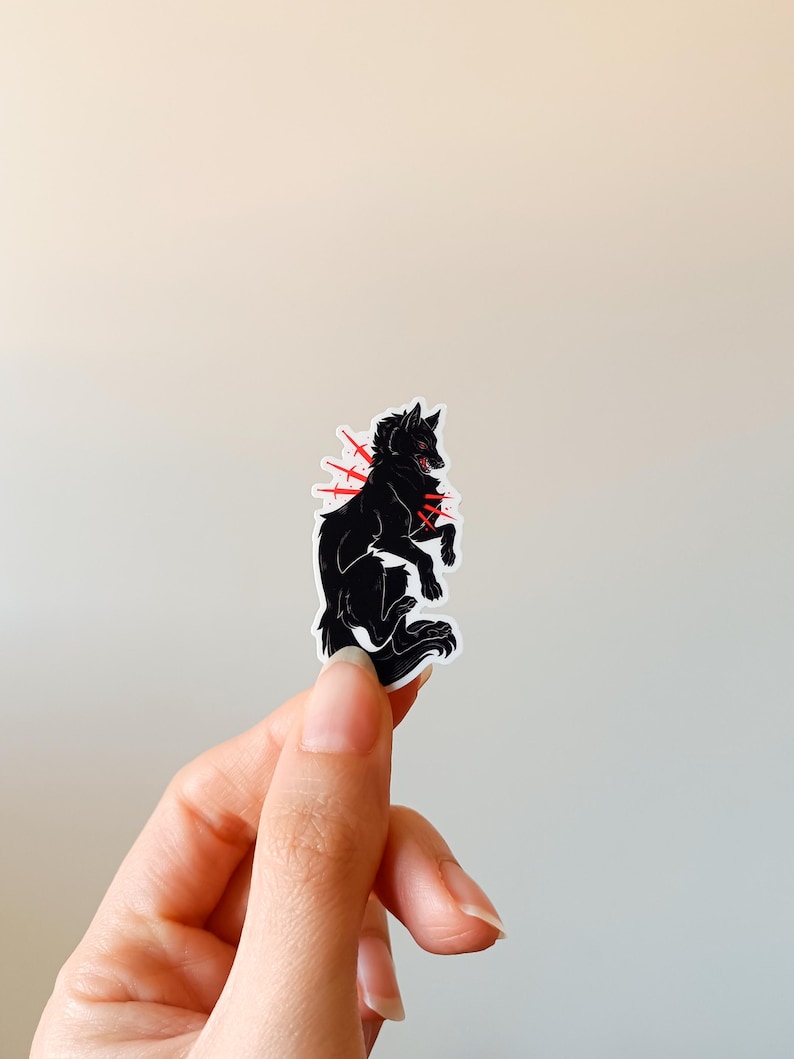 Puede incluir: Pegatina de lobo negro con detalles en rojo. El lobo se representa en una pose din&aacute;mica, con l&iacute;neas rojas que irradian de su cuerpo. La pegatina est&aacute; sujeta por una mano sobre un fondo neutro. Ilustraci&oacute;n gr&aacute;fica.
