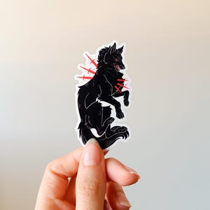 Puede incluir: Una pegatina de lobo negro con detalles en rojo. El lobo est&aacute; representado en una pose din&aacute;mica, con la boca abierta y los ojos rojos. Explosiones rojas emanan del pecho del lobo, a&ntilde;adiendo un efecto dram&aacute;tico. La pegatina se sostiene sobre un fondo neutro.