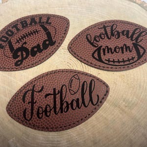 May include: Three brown leather football-shaped ornaments with black lettering. The ornaments read "Football Dad", "Football Mom", and "Football".