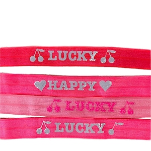 Op de afbeelding: Vier roze haarbandjes met zilveren glitterletters. De haarbandjes zeggen "LUCKY" met een kersendesign, "HAPPY" met een hartjesdesign, "LUCKY" met een muzieknootdesign en "LUCKY" met een kersendesign.