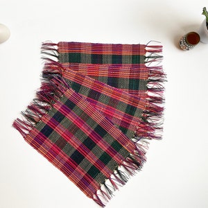 May include: Set of three woven placemats with a plaid pattern in shades of pink, green, and black. The placemats have fringed edges.