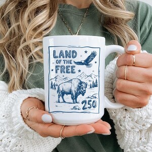May include: White ceramic mug with a blue graphic design featuring the text "LAND OF THE FREE", a flying eagle, a bison, and the number "250". The mug has a large handle and is held by a person.