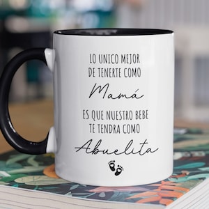 May include: White ceramic mug with a black handle and black rim. The mug has black text that reads "Lo unico mejor de tenerte como Mama es que nuestro bebe te tendra como Abuelita" with two black footprints below the text.