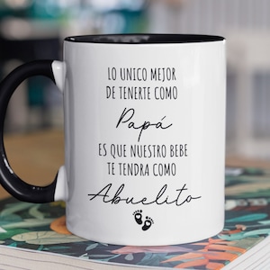 May include: White ceramic mug with a black handle and black rim. The mug has black text that reads "LO UNICO MEJOR DE TENERTE COMO Papá ES QUE NUESTRO BEBE TE TENDRA COMO Abuelito" with two small footprints below the text.