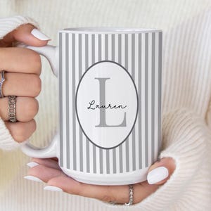 May include: White ceramic mug with vertical gray stripes. An oval in the center features the name "Lauren" and the letter "L" in gray. The mug has a white handle and is held by a person wearing a white sweater.