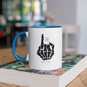 May include: White ceramic mug with a blue rim and handle. The mug features a black skeleton hand with a gold ring on the index finger. The text "MR." is above the hand and "EST. 2024" is below the hand.