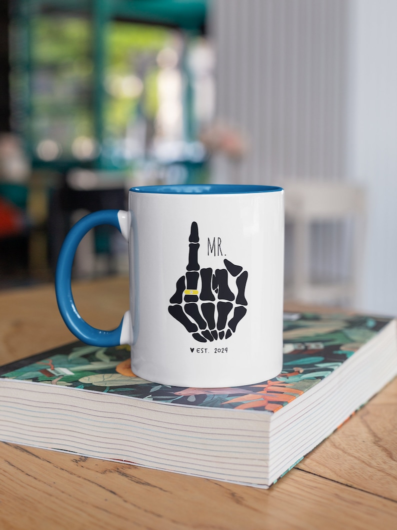 May include: White ceramic mug with a blue rim and handle. The mug features a black skeleton hand with a gold ring on the index finger. The text "MR." is above the hand and "EST. 2024" is below the hand.