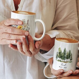 Pode incluir: Duas canecas de cer&acirc;mica branca com al&ccedil;as. Uma caneca tem um desenho floral com flores amarelas, brancas e verdes. A outra caneca tem um desenho de aquarela de uma floresta com &aacute;rvores verdes e o texto "Dad est. 2021".