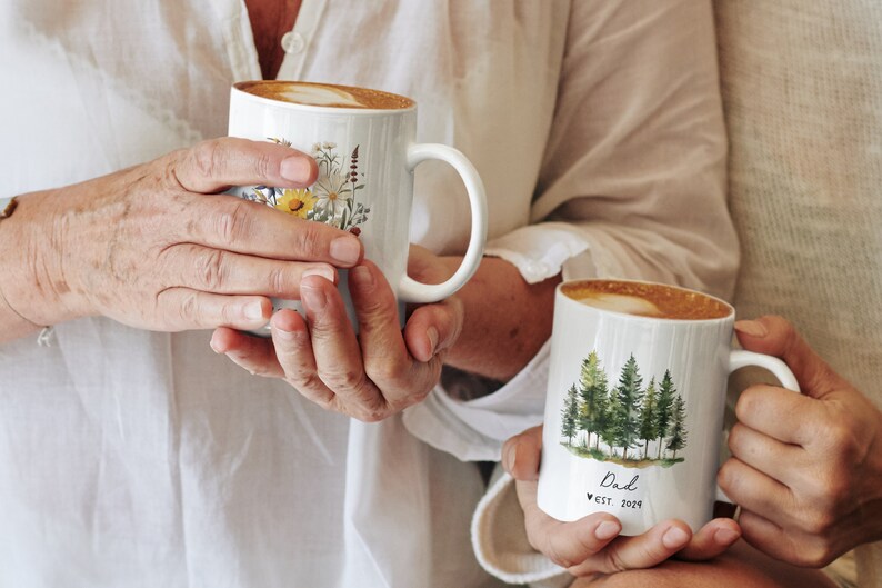 Pode incluir: Duas canecas de cer&acirc;mica branca com al&ccedil;as. Uma caneca tem um desenho floral com flores amarelas, brancas e verdes. A outra caneca tem um desenho de aquarela de uma floresta com &aacute;rvores verdes e o texto "Dad est. 2021".