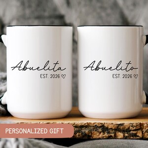 May include: Two white ceramic mugs with black handles and black rims. The mug on the left says "Abuelita Est. 2026" and the mug on the right says "Abuelito Est. 2026".
