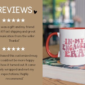 May include: White ceramic coffee mug with a red rim and handle. The mug has a pink and white graphic that says "In My Engaged Era" with a ring and stars. The mug is sitting on a stack of books.