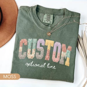 Personalized Comfort Colors moss shirt with custom floral patchwork name text, folded flat lay with boho hat and necklace, perfect grandma, nana, mimi, or mama birthday gift tee.