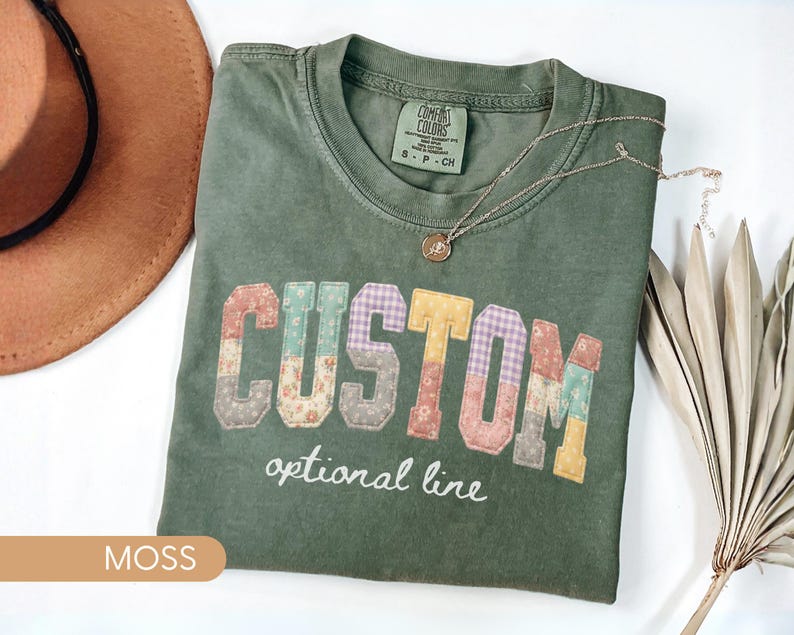 Personalized Comfort Colors moss shirt with custom floral patchwork name text, folded flat lay with boho hat and necklace, perfect grandma, nana, mimi, or mama birthday gift tee.