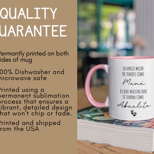 May include: White ceramic mug with a pink rim and handle. The mug has black text that reads "Lo unico mejor de tenerte como Mama es que nuestro bebe te tendra como Abuelita" with a pink footprint graphic. The mug is sitting on a stack of books.