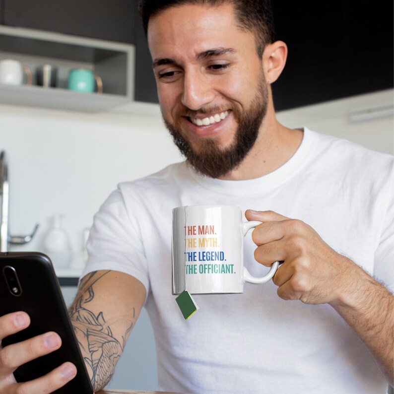 K&ouml;nnte beinhalten: Eine wei&szlig;e Keramiktasse mit dem Text "The Man. The Myth. The Legend. The Officiant." in bunten Farben. Eine Person h&auml;lt die Tasse und ein Smartphone.