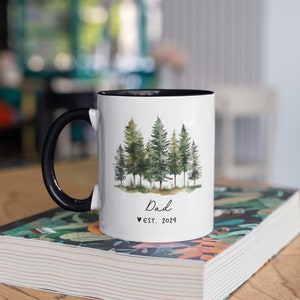 Pode incluir: Caneca de cer&acirc;mica branca com al&ccedil;a e borda pretas. A caneca apresenta uma ilustra&ccedil;&atilde;o em aquarela de uma floresta com &aacute;rvores verdes. O texto "Dad &hearts; EST. 2024" est&aacute; impresso na caneca.