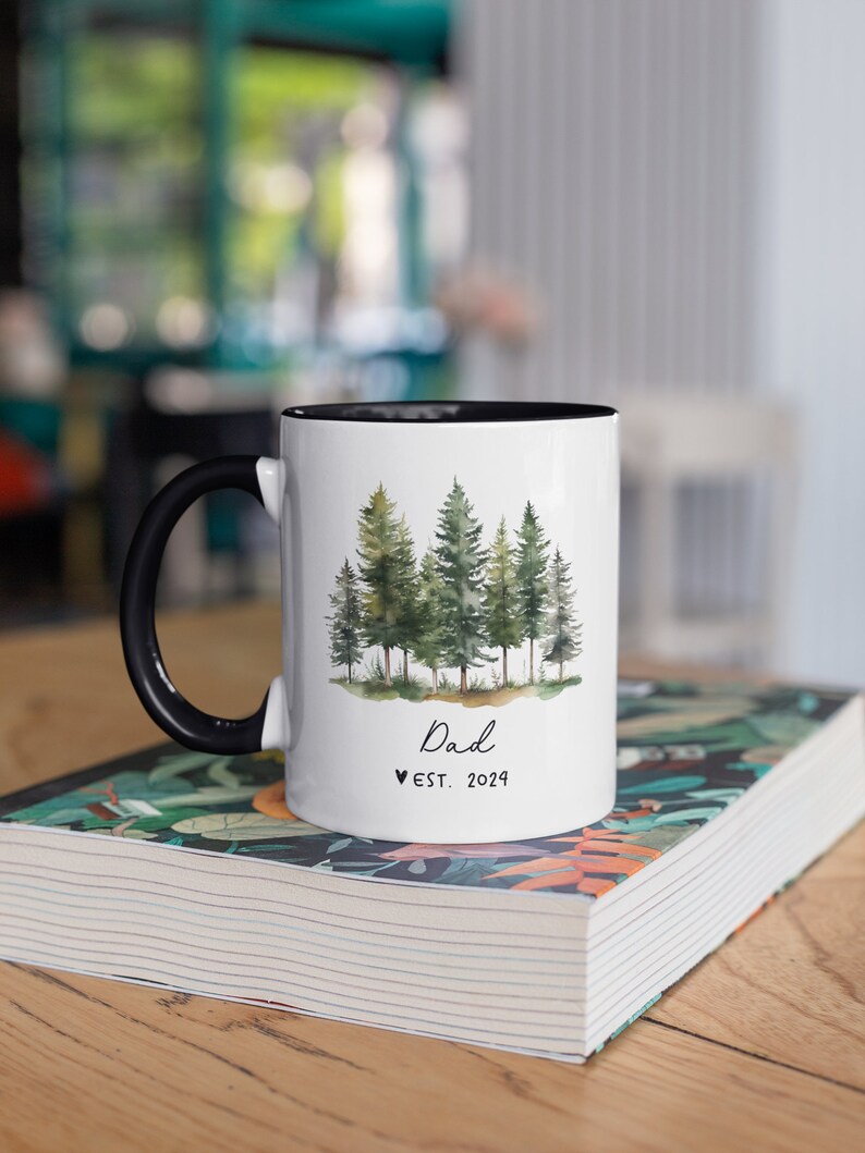Pode incluir: Caneca de cer&acirc;mica branca com al&ccedil;a e borda pretas. A caneca apresenta uma ilustra&ccedil;&atilde;o em aquarela de uma floresta com &aacute;rvores verdes. O texto "Dad &hearts; EST. 2024" est&aacute; impresso na caneca.