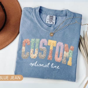 Personalized Comfort Colors blue jean shirt with custom floral patchwork name text, folded flat lay with boho hat and necklace, perfect grandma, nana, mimi, or mama birthday gift tee.