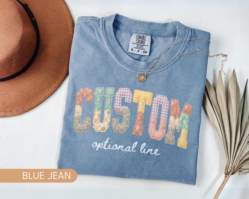 Personalized Comfort Colors blue jean shirt with custom floral patchwork name text, folded flat lay with boho hat and necklace, perfect grandma, nana, mimi, or mama birthday gift tee.