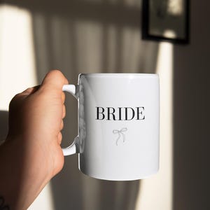 May include: White ceramic mug with a black inscription that reads "BRIDE" and a small black bow.