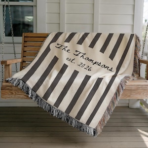May include: A cream-colored throw blanket with vertical gray stripes is draped over a wooden swing. The blanket features the embroidered text "The Thompsons est. 2026" in a decorative font, and has fringed edges.