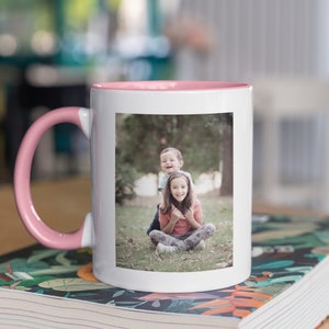 May include: White ceramic mug with a pink interior and handle. The mug displays a rectangular photo of two children. The mug is on a book with a colourful floral design.