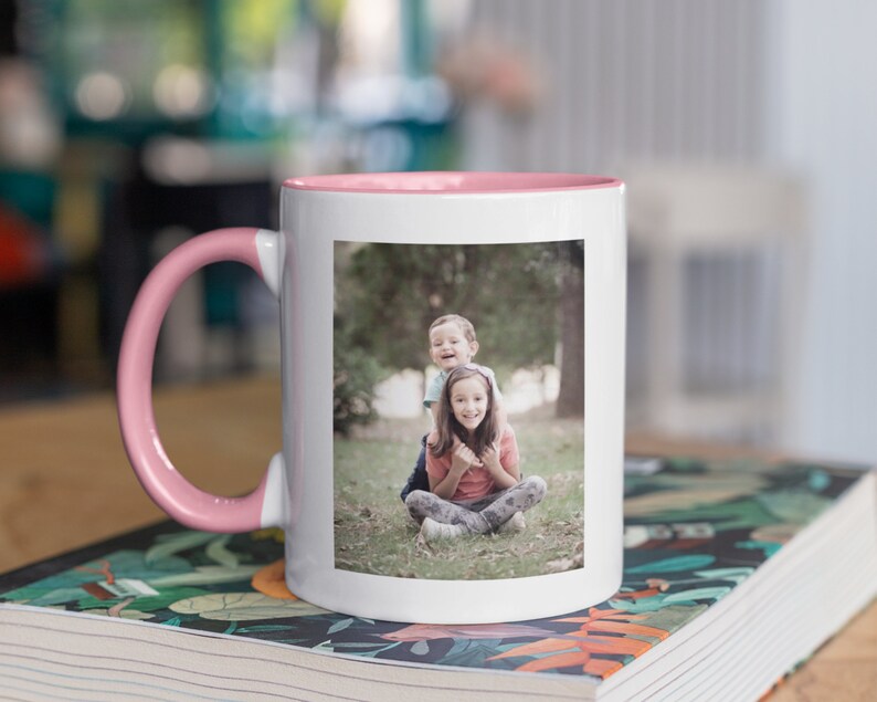 May include: White ceramic mug with a pink interior and handle. The mug displays a rectangular photo of two children. The mug is on a book with a colourful floral design.