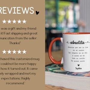 Pode incluir: Caneca de cer&acirc;mica branca com interior e al&ccedil;a vermelhos. A caneca apresenta a palavra "abuelita" em escrita cursiva, juntamente com um poema em espanhol e o nome "Sophia". A caneca est&aacute; em um livro.