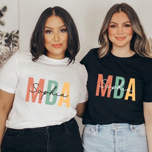 Op de afbeelding: Twee mensen dragen T-shirts met de tekst "MBA Sophia" in kleurrijke letters. De persoon links draagt een wit T-shirt en de persoon rechts draagt een zwart T-shirt.