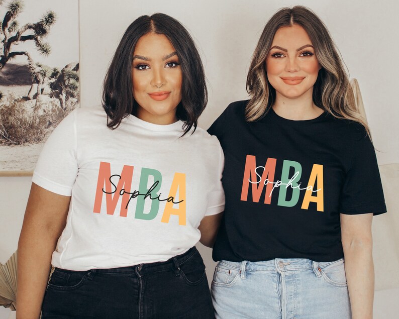 Op de afbeelding: Twee mensen dragen T-shirts met de tekst "MBA Sophia" in kleurrijke letters. De persoon links draagt een wit T-shirt en de persoon rechts draagt een zwart T-shirt.