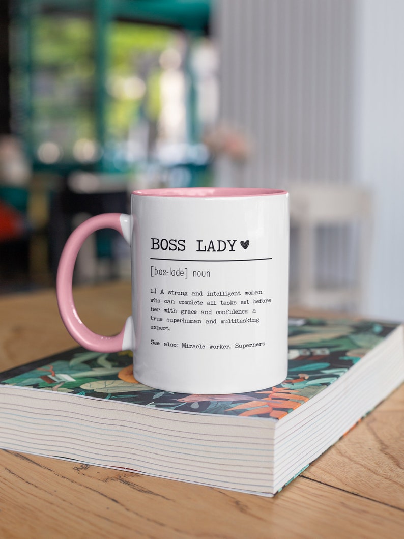 May include: White ceramic mug with a pink rim and handle. The mug has black text that reads "BOSS LADY [bos-lade] noun 1) A strong and intelligent woman who can complete all tasks set before her with grace and confidence a true superhuman and multitasking expert. See also: Miracle worker, Superhero"