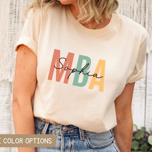 Op de afbeelding: Een cr&egrave;mekleurig T-shirt met de tekst "MBA" in kleurrijke letters en de naam "Sophia" eronder in cursief.