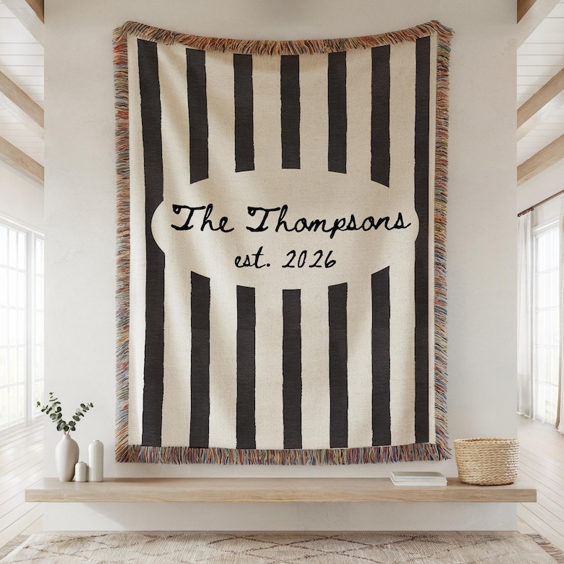 May include: A large, cream-colored throw blanket with vertical black stripes. The blanket features a cream-colored oval with the text "The Thompsons est. 2026" in black script. The edges have a fringed trim.