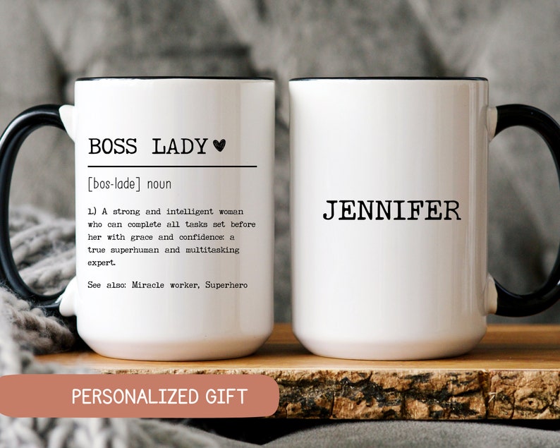 May include: Two white ceramic mugs with black handles and black rims. The mug on the left has the text "BOSS LADY [bos-lade] noun 1.) A strong and intelligent woman who can complete all tasks set before her with grace and confidence: a true superhuman and multitasking expert. See also: Miracle worker, Superhero PERSONALIZED GIFT" The mug on the right has the text "JENNIFER".