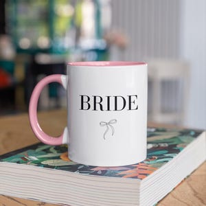 May include: White ceramic mug with a pink rim and handle. The mug has the word "BRIDE" printed in black with a small bow underneath.