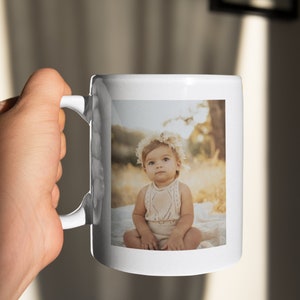 May include: White ceramic mug with a photograph of a baby wearing a floral crown and a knitted outfit. The mug has a curved handle and a glossy finish. The photograph's background is a blurred outdoor scene.