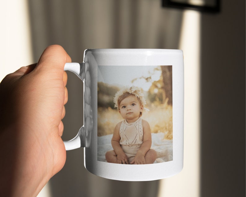 May include: White ceramic mug with a photograph of a baby wearing a floral crown and a knitted outfit. The mug has a curved handle and a glossy finish. The photograph's background is a blurred outdoor scene.