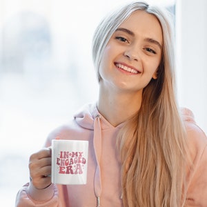 May include: A white ceramic mug with the text "In My Engaged Era" in pink lettering. The mug has a pink ring around the top and a pink ring around the bottom. A woman is holding the mug in her right hand.