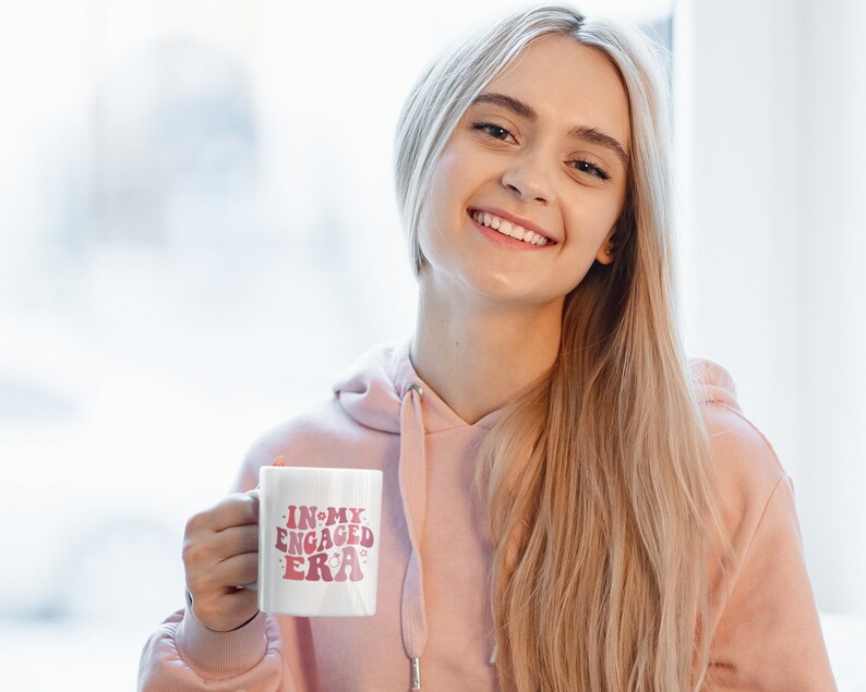 May include: A white ceramic mug with the text "In My Engaged Era" in pink lettering. The mug has a pink ring around the top and a pink ring around the bottom. A woman is holding the mug in her right hand.