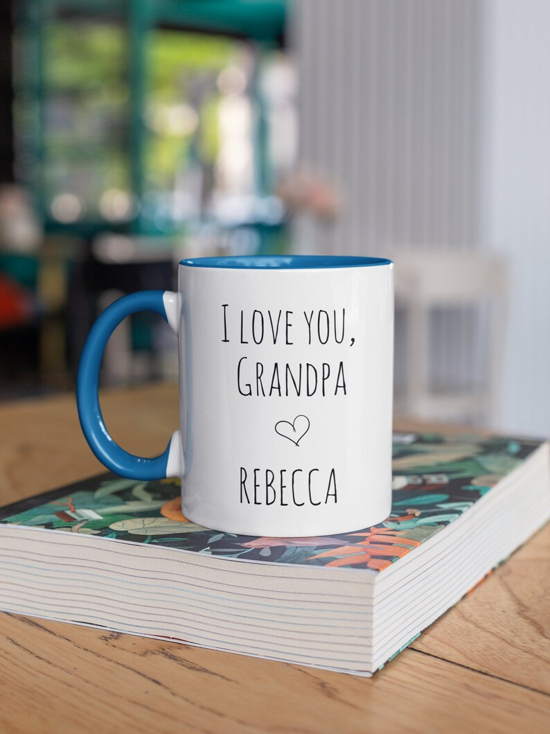 May include: White ceramic mug with a blue interior and handle. The mug has the text "I LOVE YOU, Grandpa" with a heart symbol and the name "Rebecca" in black lettering. The mug is on a stack of books.