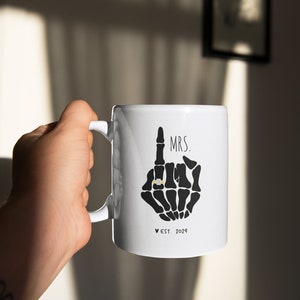 May include: White ceramic mug featuring a black skeleton hand design. The hand is making a middle finger gesture with a gold ring on the index finger. The text "MRS. EST. 2024" is printed below the hand.