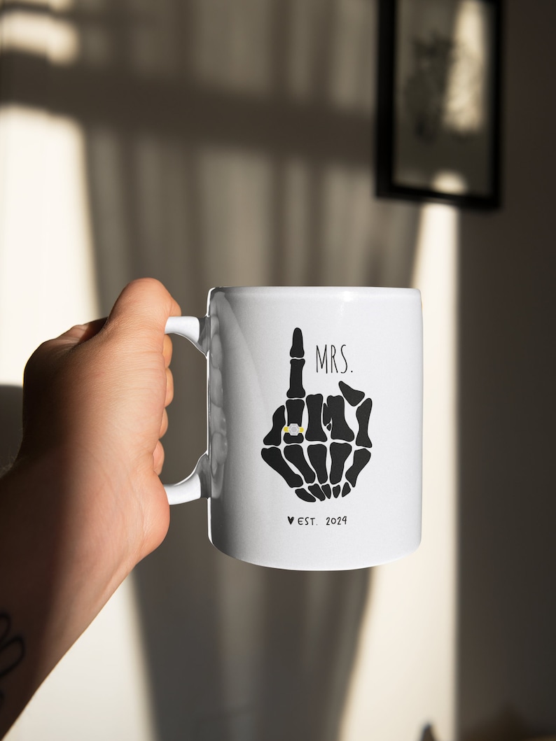 May include: White ceramic mug featuring a black skeleton hand design. The hand is making a middle finger gesture with a gold ring on the index finger. The text "MRS. EST. 2024" is printed below the hand.