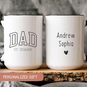 May include: Two white ceramic mugs with black handles and rims. One mug has the word "DAD" and "EST. 2023&2024" printed in black. The other mug has the names "Andrew Sophia" and a black heart. The mugs are on a wooden surface.
