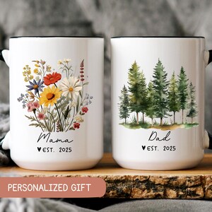 Pode incluir: Duas canecas de cer&acirc;mica branca com al&ccedil;as pretas. A caneca &agrave; esquerda tem um desenho floral e o texto "Mama Est. 2025". A caneca &agrave; direita tem uma cena de floresta em aquarela e o texto "Dad Est. 2025".